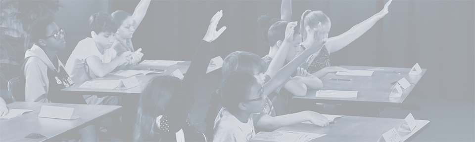 A group of children seated at desks. Some are raising their hands.